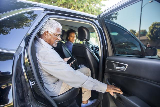 Older man closing the passenger side door to a car. An adult woman is driving.