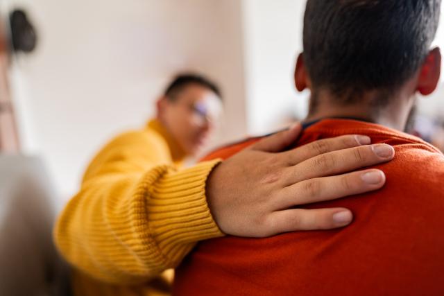 A person comforting a man by placing their hand on his shoulder.