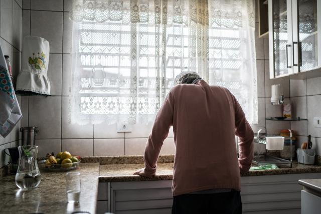 An older adult stands with their back to the camera, leaning on a kitchen counter with head bowed, in a sunlit kitchen with lace curtains, tiled walls, and a bowl of fruit on the counter.