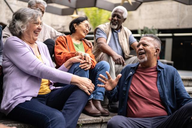 A diverse group of older adults sit together outdoors on steps, laughing and chatting animatedly with one another.