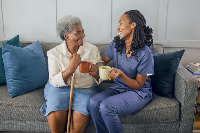 An older woman with a cane and a caregiver in navy scrubs sit together on a couch, laughing while holding mugs of coffee.