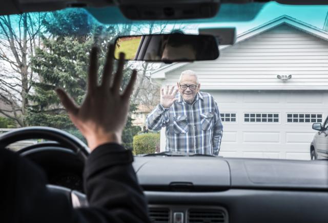 Viewed through a car windshield, a smiling older man in a plaid shirt waves goodbye from a driveway in front of a garage, while a person in the driver's seat waves back.