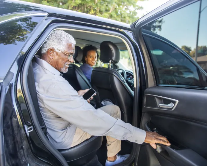 Older man closing the passenger side door to a car. An adult woman is driving.