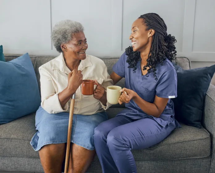 An older woman with a cane and a caregiver in navy scrubs sit together on a couch, laughing while holding mugs of coffee.
